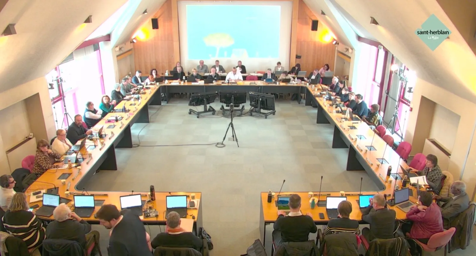 Plan large de la salle du conseil de Saint-Herblain : les élus installés en U, le logo de la ville à l'écran